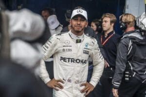 Lewis Hamilton in his white Mercedes race overalls and a hat on his head, standing with his hands on his hips looking at the camera.